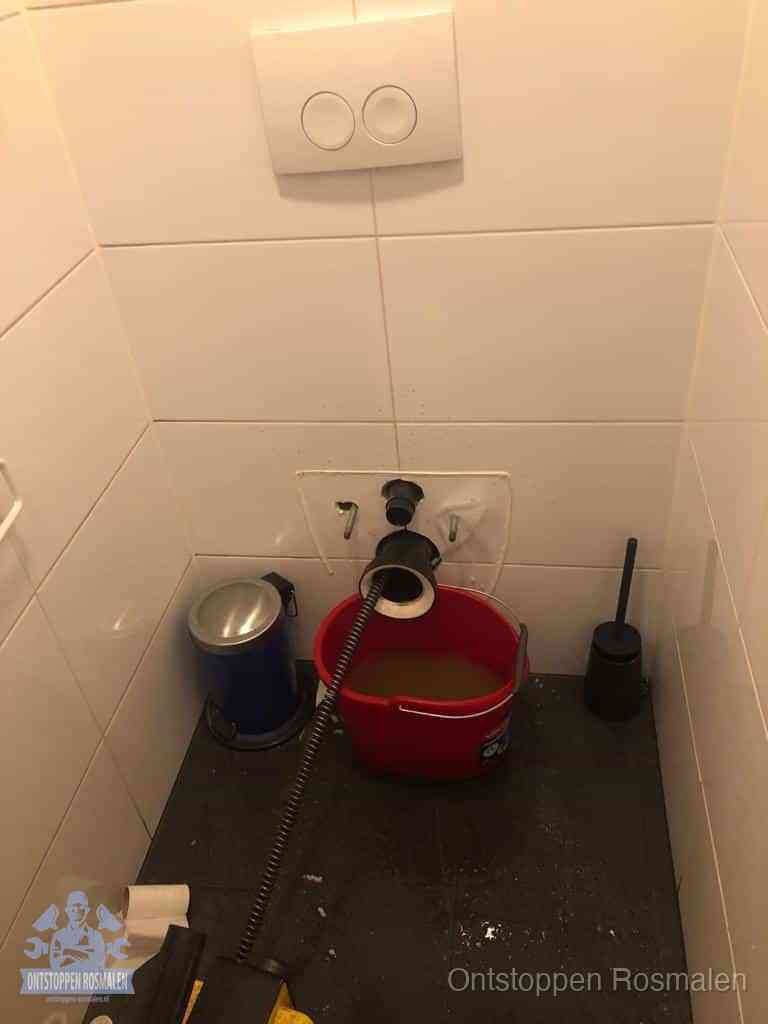 Een ontstopte wand-wc met verwijderde toiletpot waar een ontstoppingsveer in de wanduitgang verdwijnt, het achterhalen waar de verstopping zit in Rosmalen
