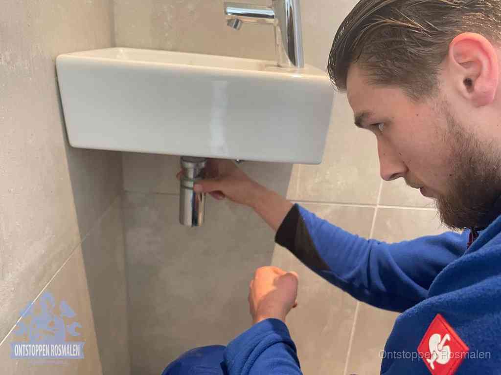 Een monteur draait met de hand de zwanenhalssifon onder een fonteinwasbak in de toiletruimte los om de bron van rioollucht in Rosmalen te vinden