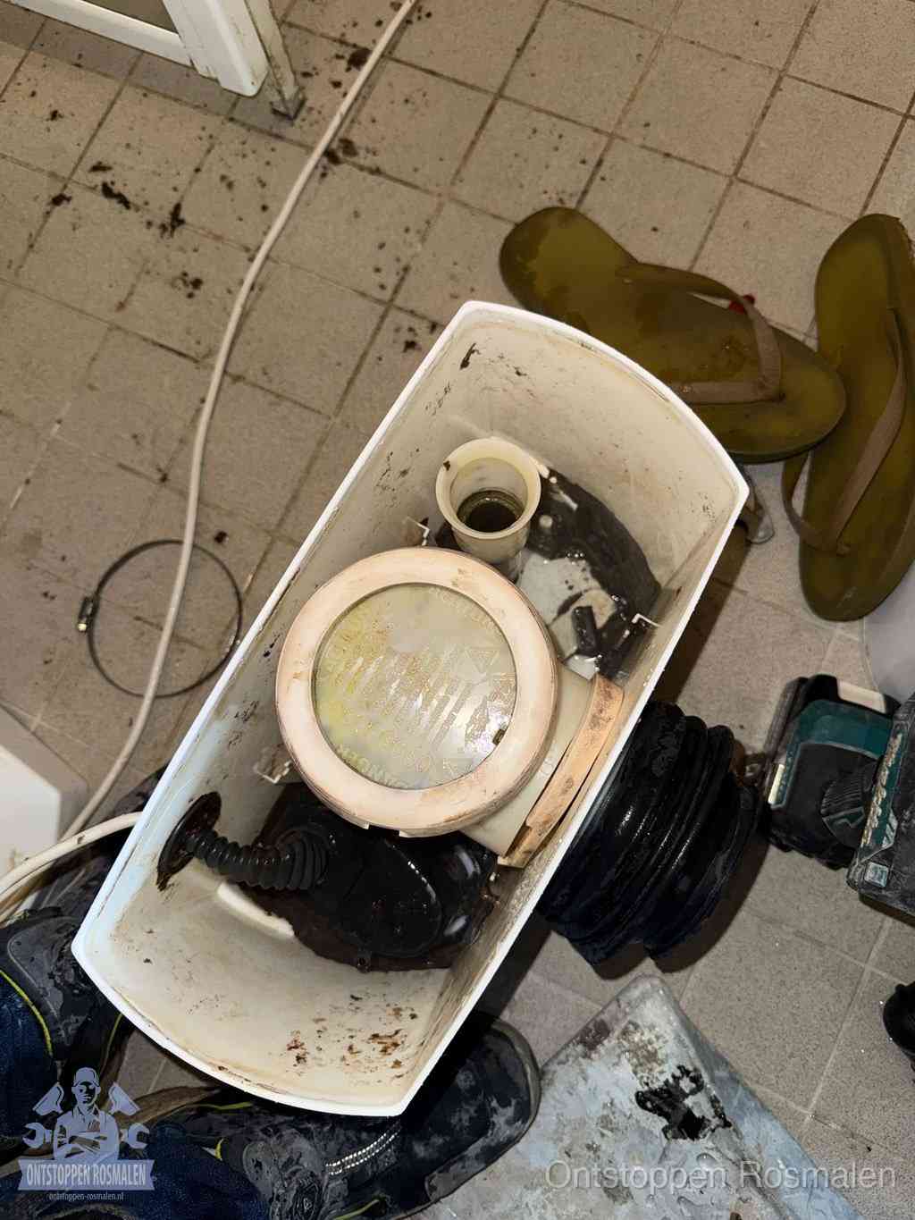 Een gedemonteerde toiletpot op de vloer met ontstoppingsveer en gereedschap ernaast, het verwijderen van een vastzittend voorwerp uit de wc in Rosmalen